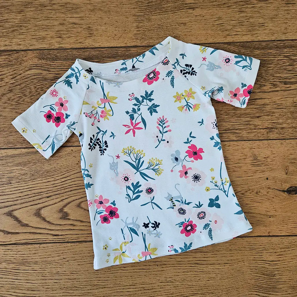 T-shirt raglan enfant Tao cousu dans un jersey blanc à motifs fleuris colorés, présenté à plat sur un sol en bois – patron de couture Tao de Kandix