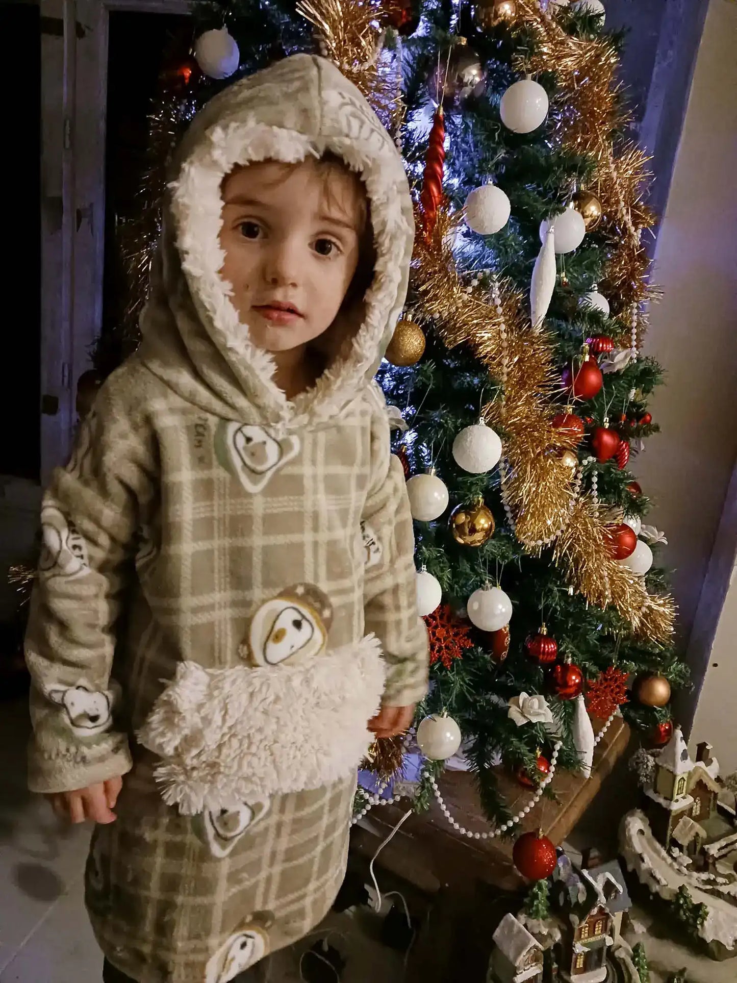 Sweat doudou enfant beige à carreaux et motifs chiens, capuche bordée de fausse fourrure, debout devant un sapin de Noël décoré - patron de couture Casimir de Kandix