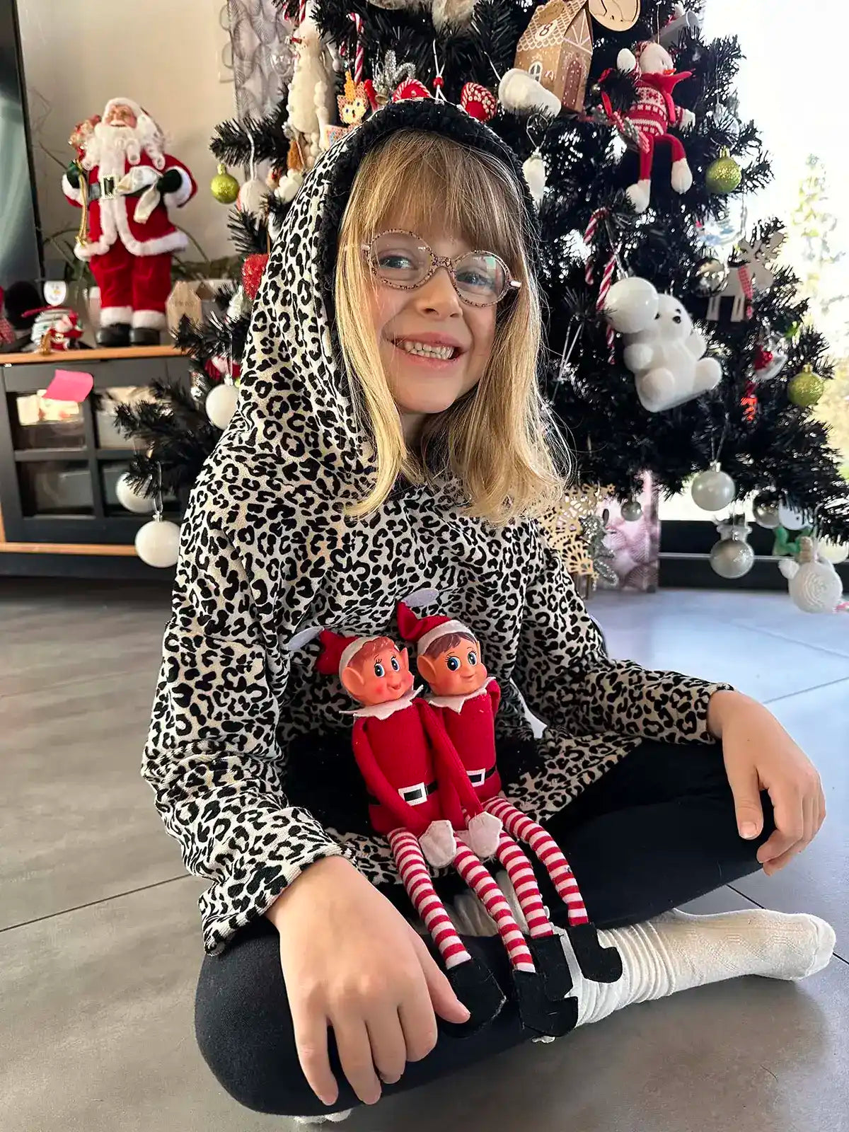 Enfant assise en tailleur devant un sapin, vêtue d’un sweat doudou léopard à capuche, deux lutins de Noël sur les genoux - patron de couture Casimir de Kandix