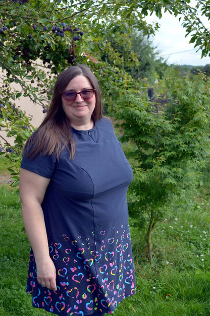 Femme souriante portant une robe bleu marine à manches courtes avec imprimé de cœurs colorés, en extérieur sous un prunier - patron de couture Gaby de Kandix