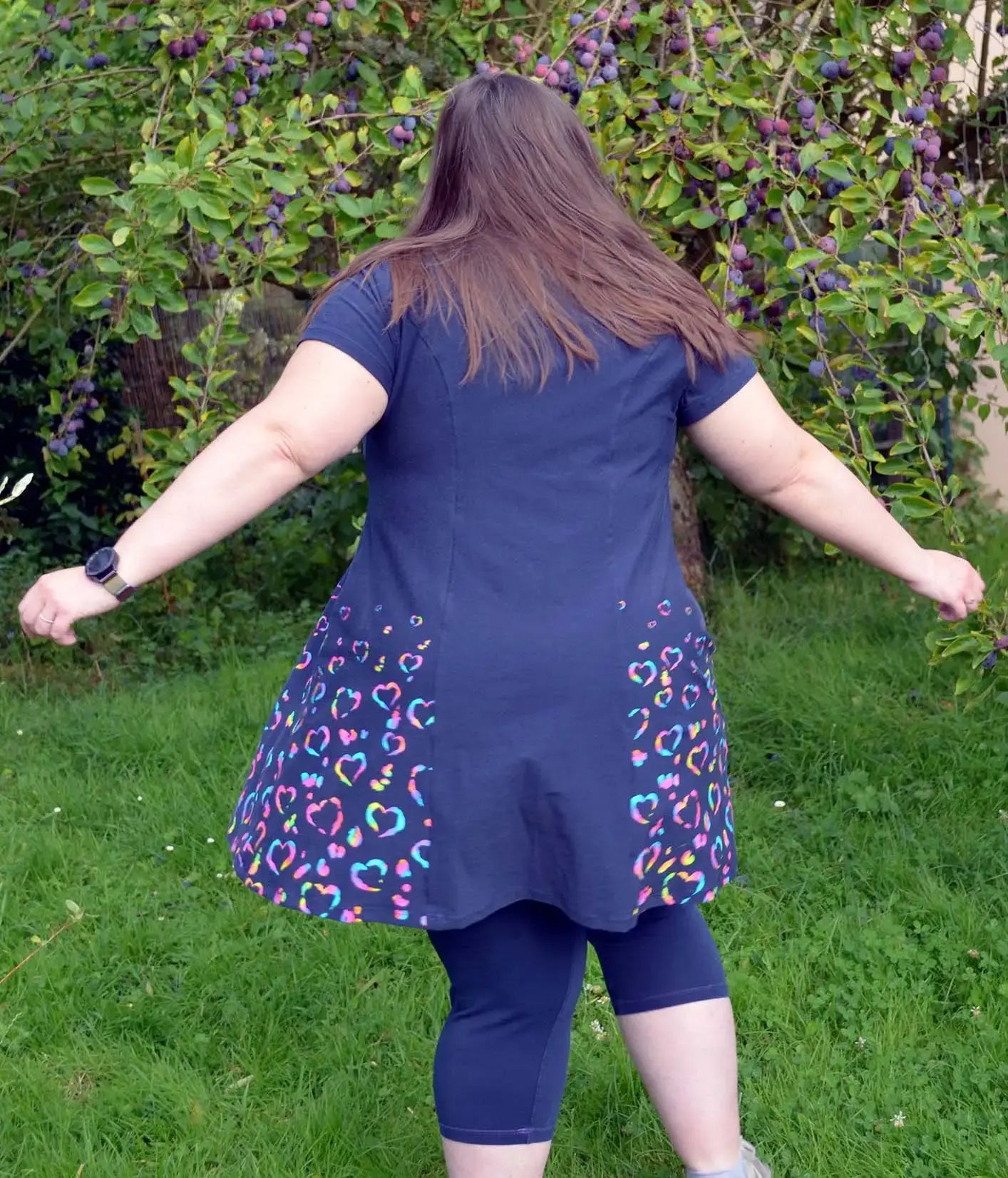 Vue de dos d'une femme tournant sur l'herbe, en robe bleu marine décorée de cœurs multicolores, dans un jardin - patron de couture Gaby de Kandix