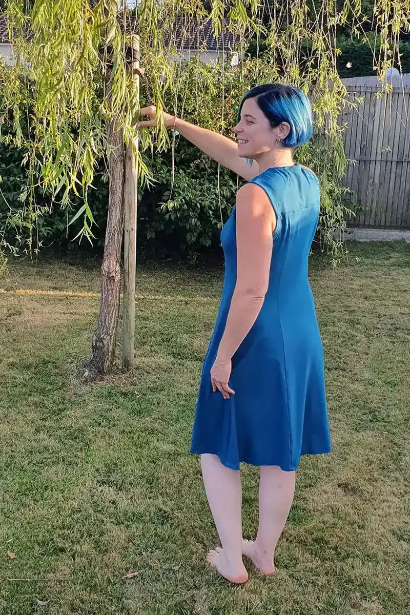 Vue de dos d'une femme en robe bleue sans manches touchant une branche de saule, dans un jardin - patron de couture Gaby de Kandix