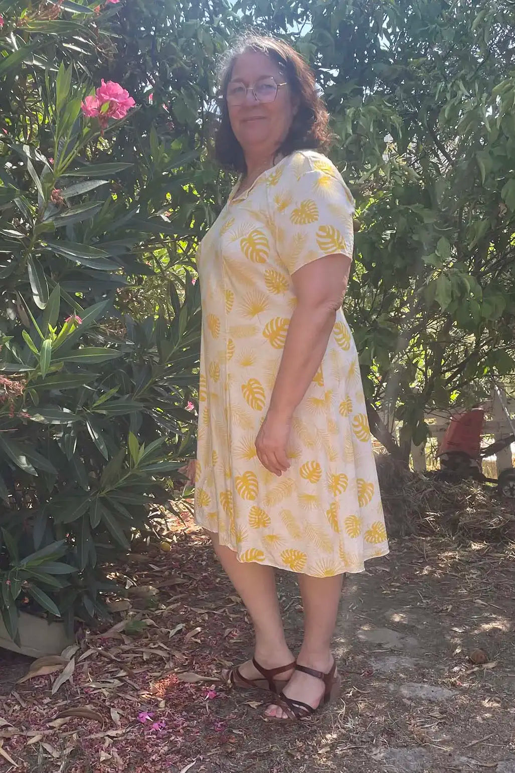 Femme mature en robe Gaby jaune à manches courtes, posant de profil dans un jardin fleuri - patron de couture Gaby de Kandix