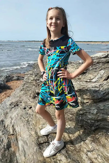 Fillette posant fièrement sur un rocher en bord de mer, vêtue d’une robe colorée à imprimé graffiti - patron de couture Gaby de Kandix