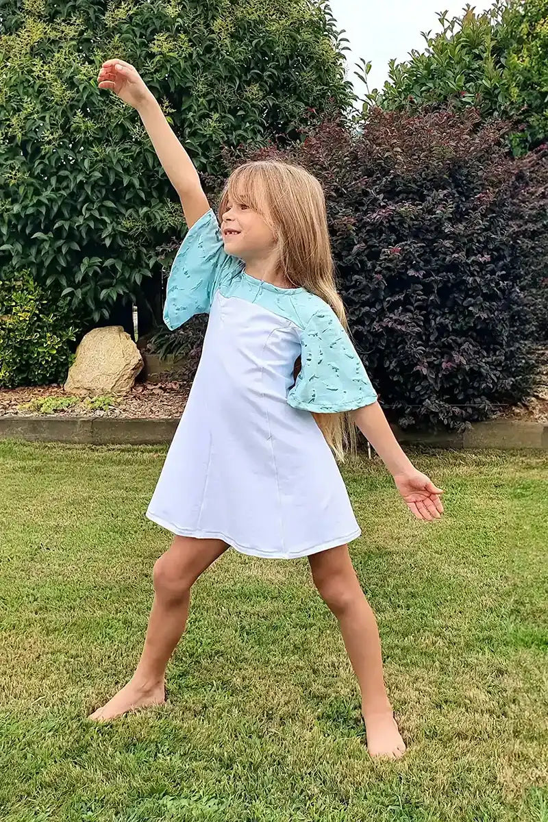 Fillette en robe turquoise à manches papillon, en pleine pose de super-héroïne dans un jardin - patron de couture Gaby de Kandix